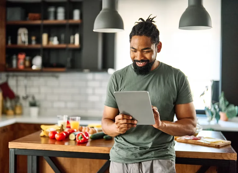 A man stands in his home kitchen using a tablet in front of a countertop containing fresh produce.