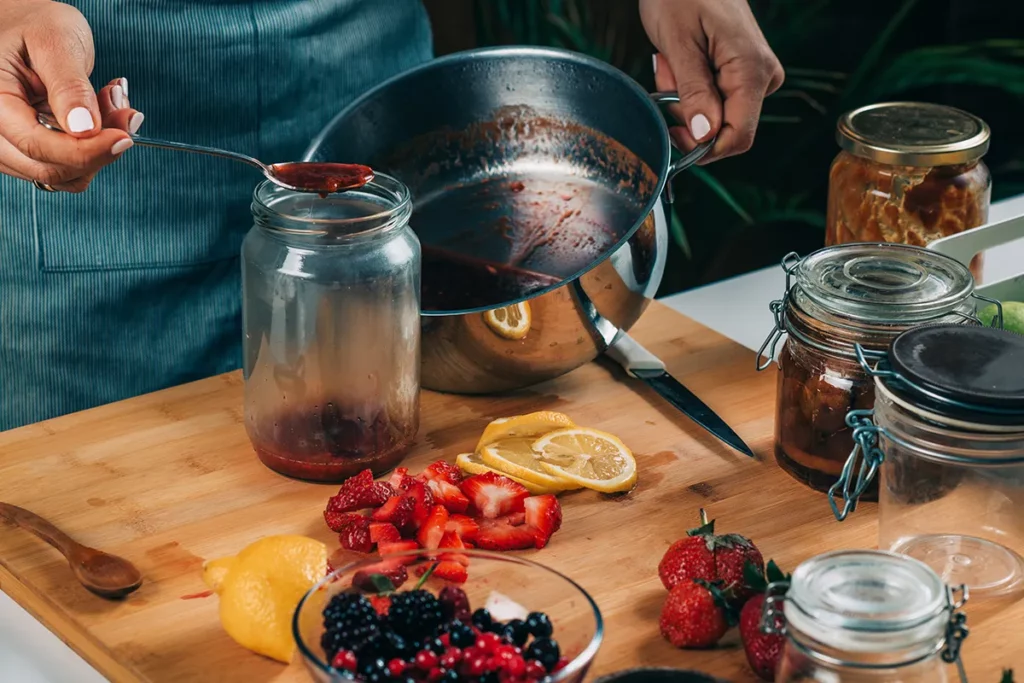 A person pouring fruit jam from a pot into a jar in a home kitchen.