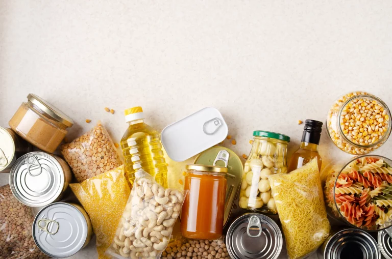 A flat lay view of non-perishable foods on a kitchen counter, including popcorn kernels, dry pasta, and canned foods.
