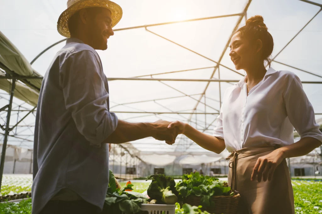 A man and a woman shake hands in front of a box of vegetables inside a greenhouse.