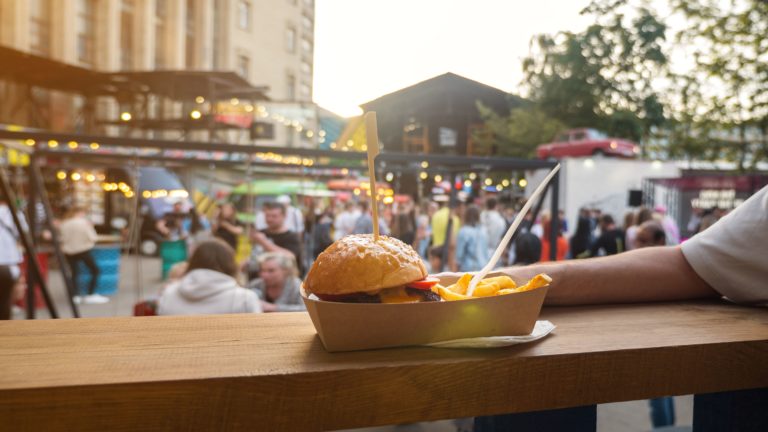 A close-up of a man holding a street food burger with French fries with a street food festival in the background.