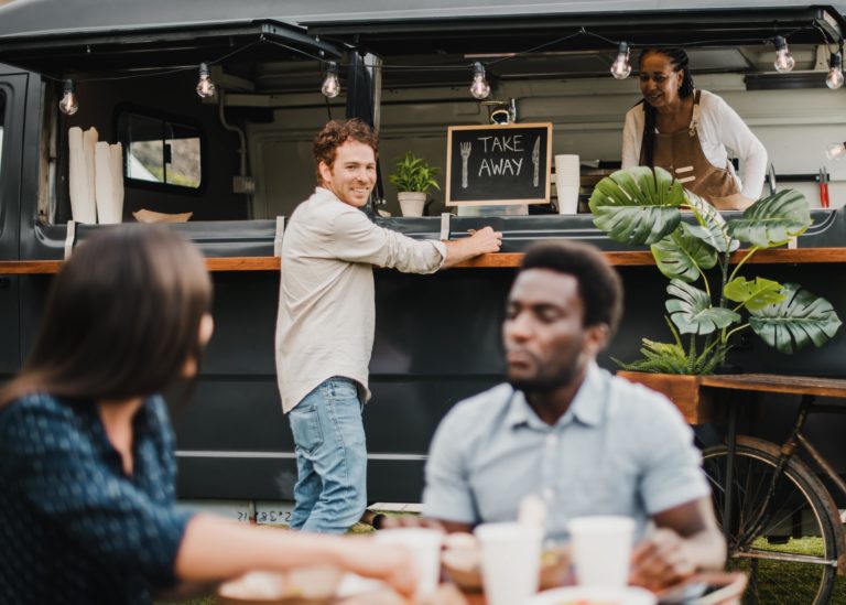 A man ordering from a black food truck looks back at his friends sitting at a table.