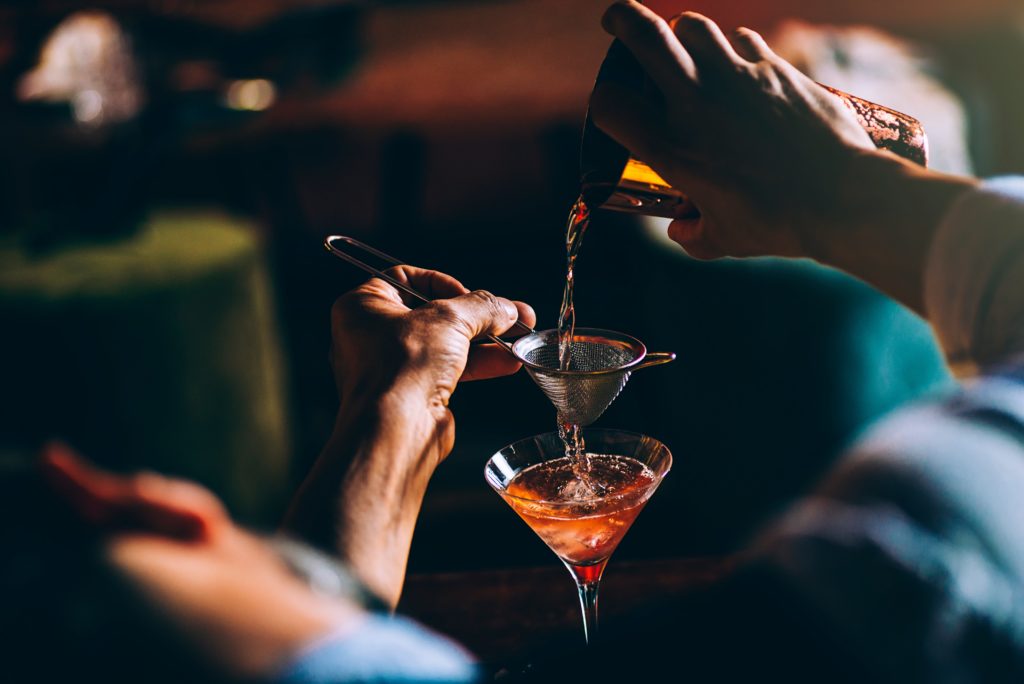 A bartender pours a cocktail through a strainer and into a glass.