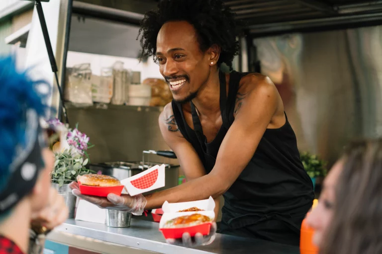 A smiling food vendor in a black apron hands two burgers to a pair of customers waiting at the window of a food truck.