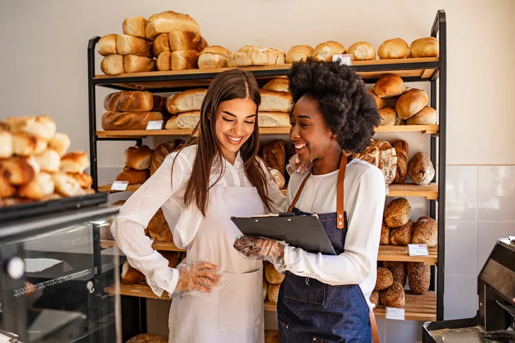 Two young women standing in front of shelves of bread in a bakery and looking at a clipboard.