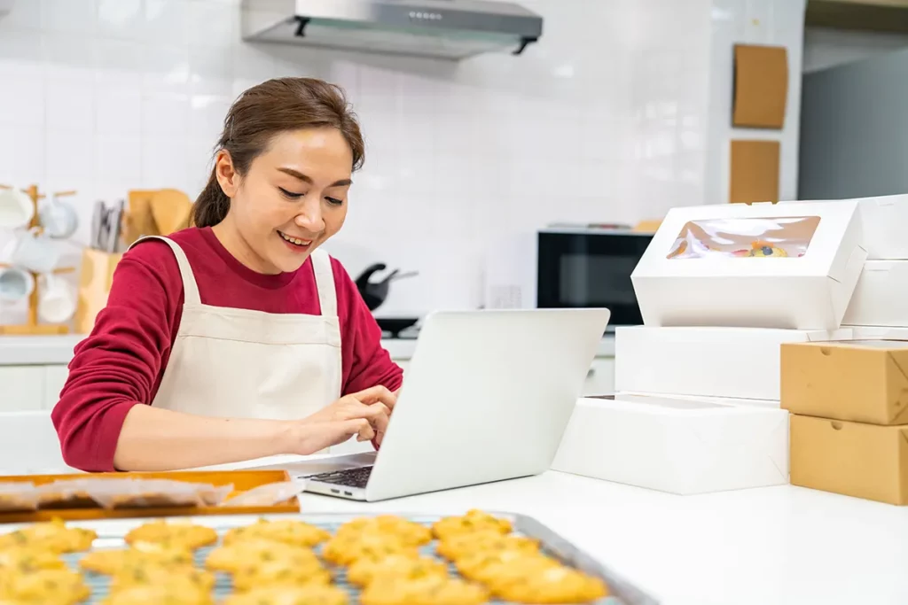 A baker types on a laptop in her kitchen next to a batch of cookies and several boxes of baked goods.