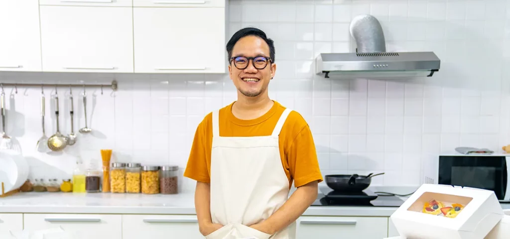 A man with glasses stands in a white kitchen in front of a countertop covered with boxes and trays of baked goods for his home baking business.