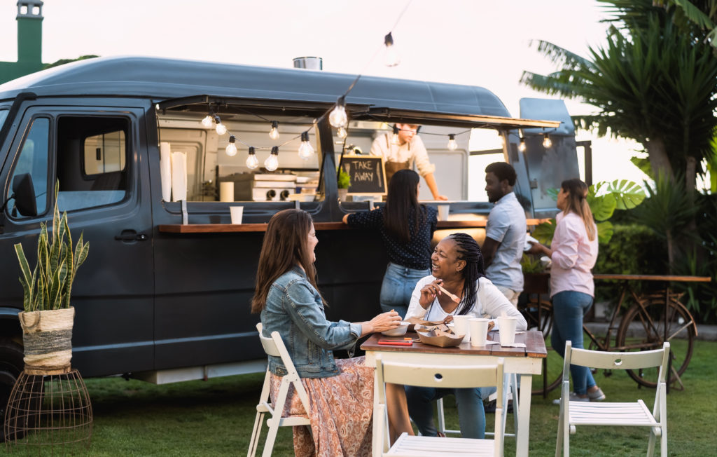 People ordering from a black food truck van while other customers eat their food at a table in the foreground.