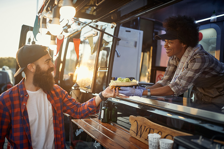 A woman working inside of a food truck hands a sandwich to a customer.