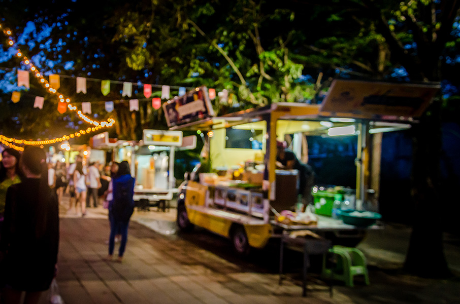 An out-of-focus shot of people walking along a path at a food truck festival in the evening.