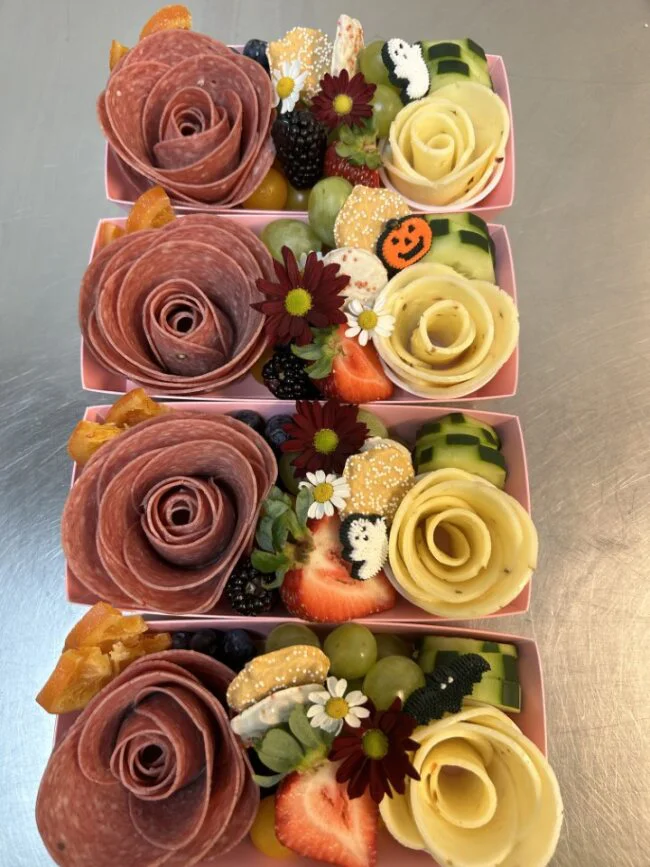 A charcuterie box from Fancy Nancy Boards containing meats and cheeses shapes like roses as well as fruit and small Halloween decorations.