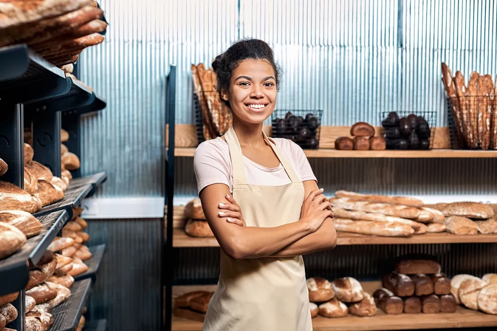 A young woman in a beige apron standing in a bakery with shelves full of bread.