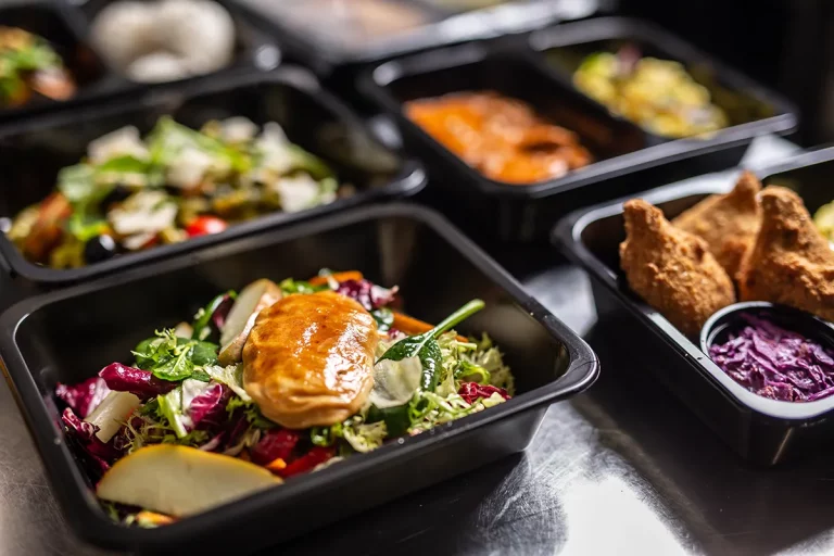 Premade meals in plastic delivery containers on a stainless steel countertop.